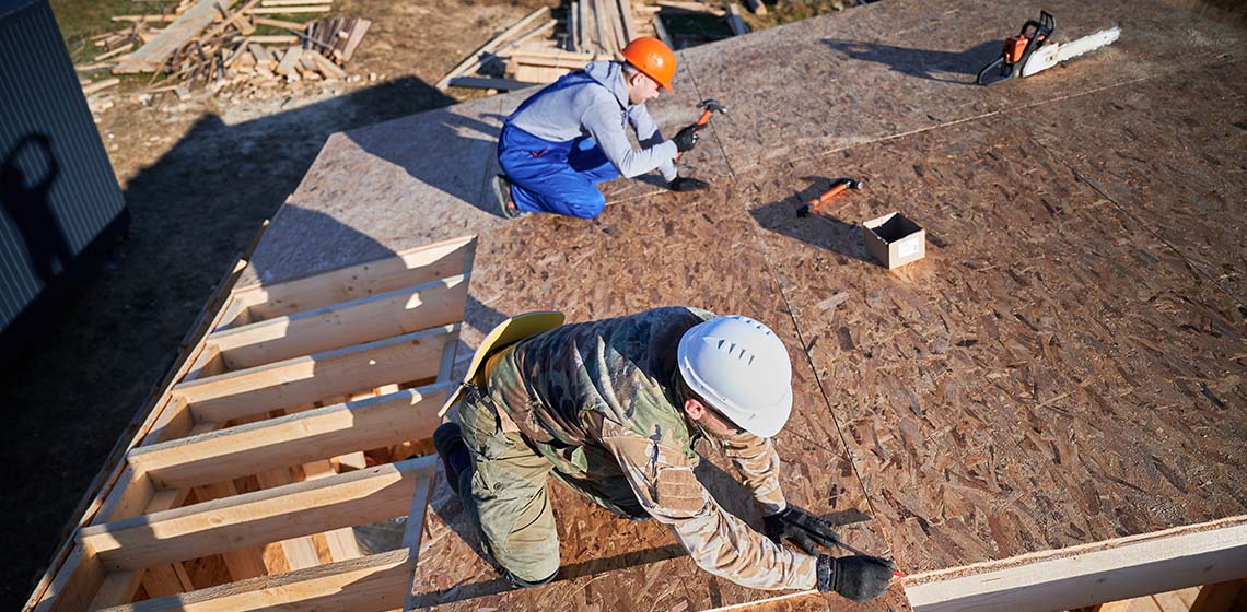 Two workers construct a roof, one kneeling on plywood and the other standing with a hammer, surrounded by tools and wooden materials.